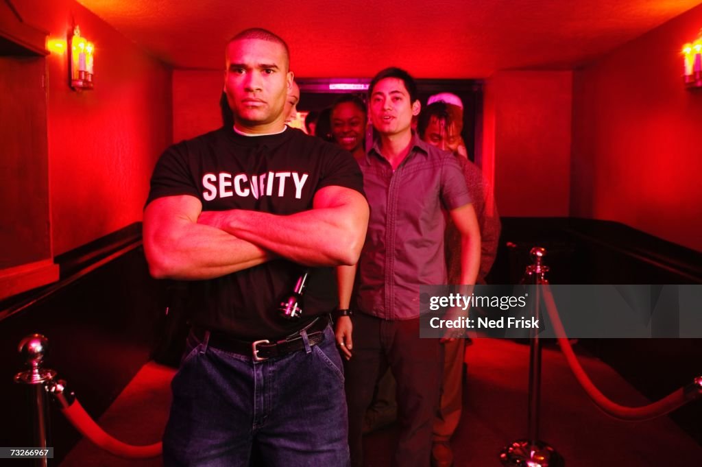 African male bouncer with arms crossed in front of line of people