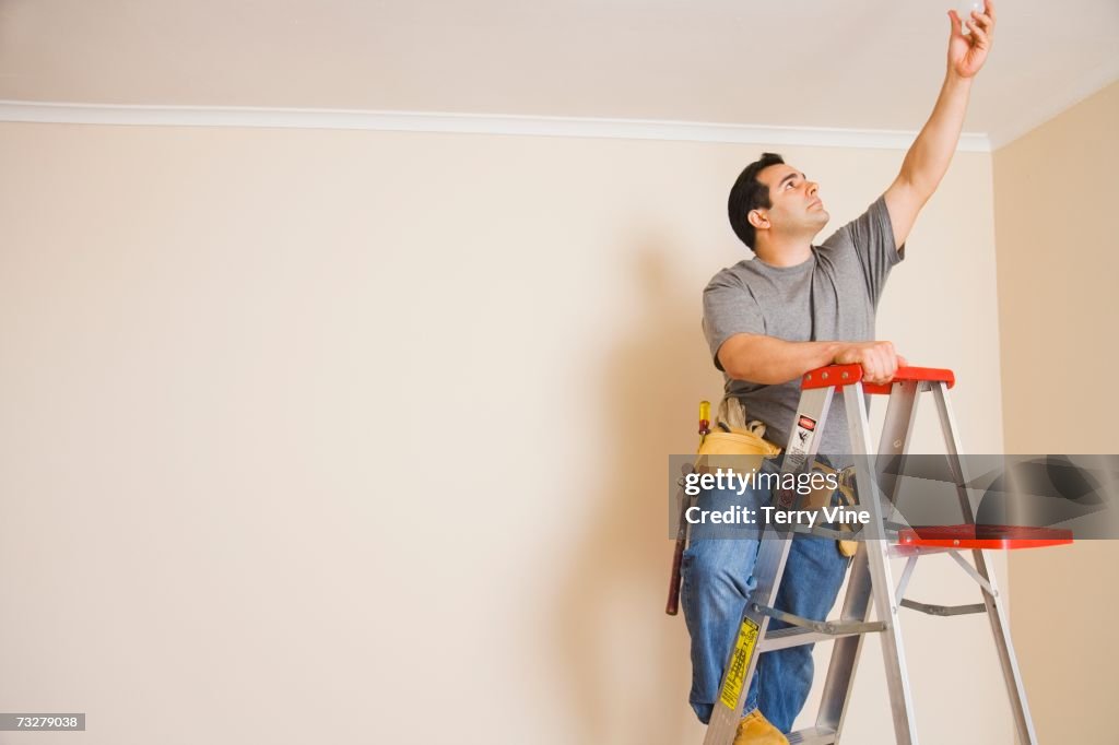 Man wearing tool belt on ladder changing light bulb