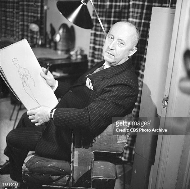 French fashion designer Christian Dior sits in a chair with a sketchpad, on which is a fashion design, for a broadcast of the CBS celebrity interview...