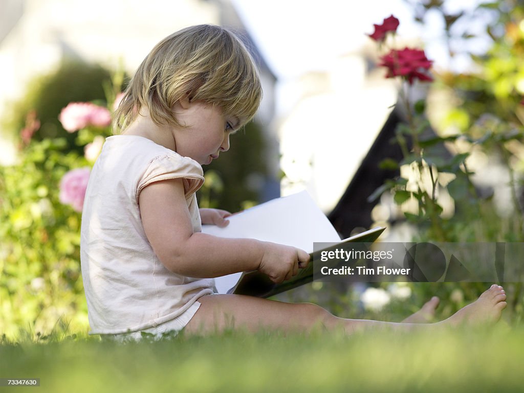 Toddler girl (18-24 months) looking at book in yard