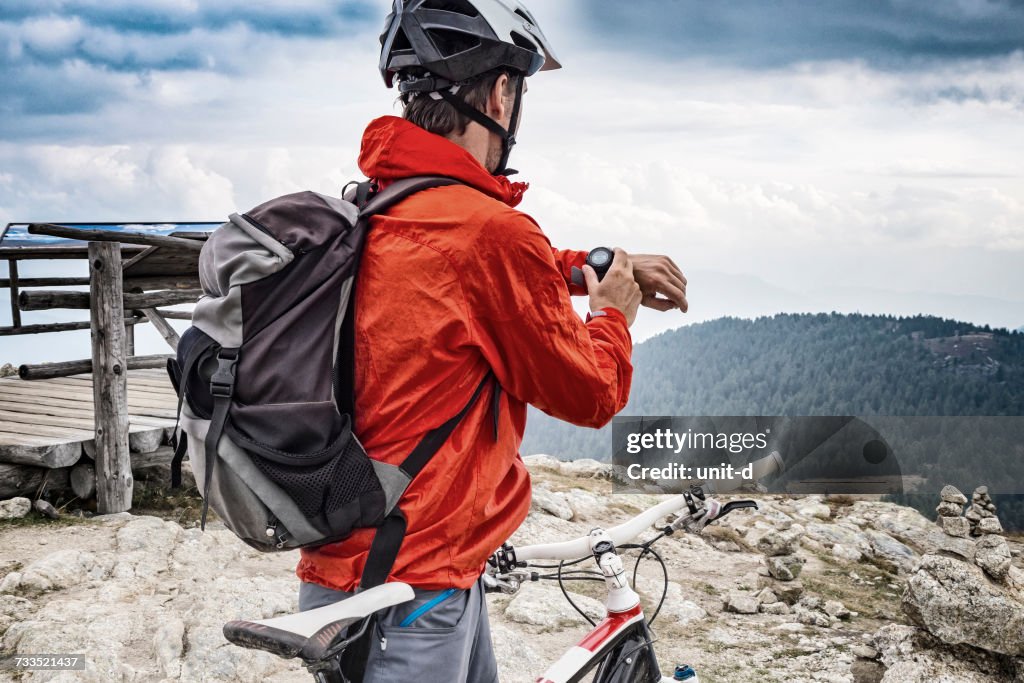 Rückansicht des Mountain Bikers, der die Smartwatch gegen bewölkten Himmel betrachtet