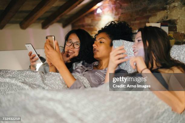 friends lying side by side in bed looking at mobile phones - sleepover bildbanksfoton och bilder