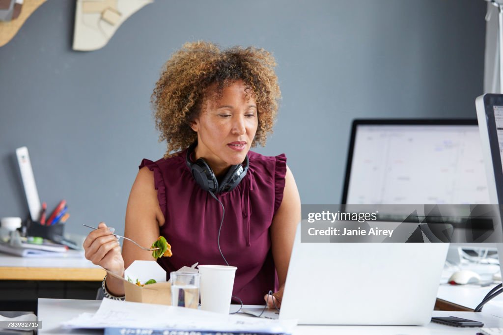 Female designer eating working lunch and looking at laptop at desk