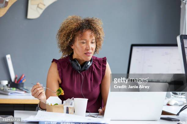 female designer eating working lunch and looking at laptop at desk - smart casual lunch stock pictures, royalty-free photos & images