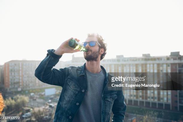 young man drinking from beer bottle at roof party - beer bottle stock-fotos und bilder