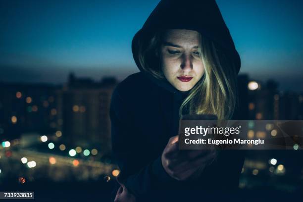 young woman sitting on rooftop at night, using smartphone, illuminating face - milan night stock pictures, royalty-free photos & images