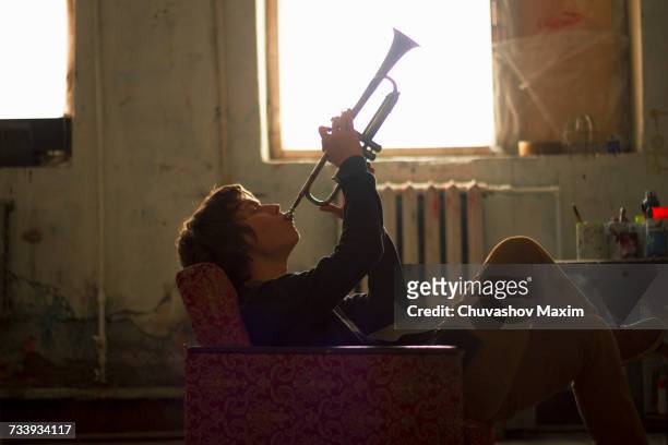 young man sitting on vintage armchair in artist studio playing trumpet - beatnik fotografías e imágenes de stock