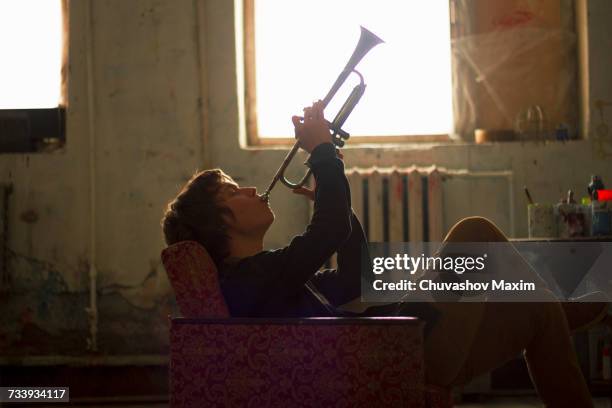 young man sitting on vintage armchair in artist studio playing trumpet - beat generation stock pictures, royalty-free photos & images