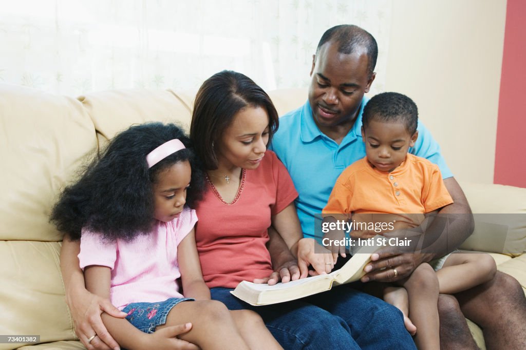 Parents with children (4-7) on sofa, reading bible, smiling