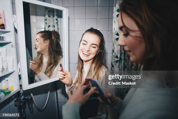 happy female friends with mobile phone and lip gloss at dorm bathroom - make up looks stock pictures, royalty-free photos & images