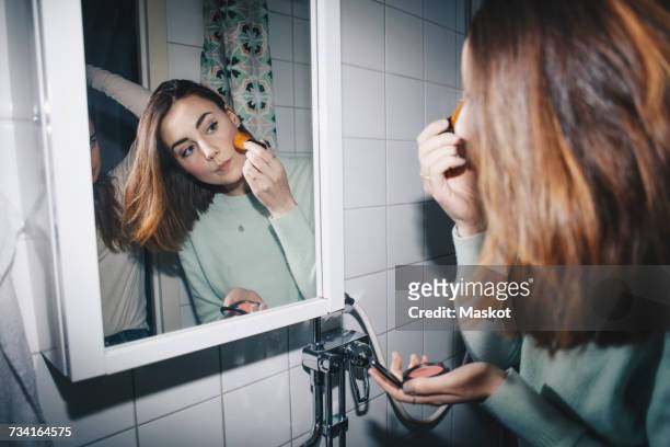 young woman applying blush looking in mirror at college dorm bathroom - applying makeup with brush fotografías e imágenes de stock