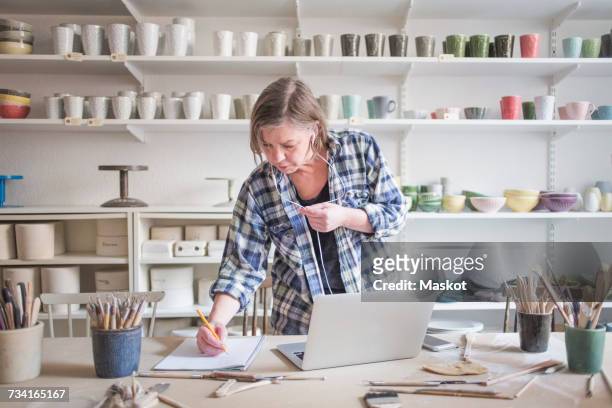mature female potter writing on paper while standing by laptop at table in workshop - 50s woman writing at table stock-fotos und bilder