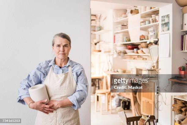 portrait of senior female potter standing with vase against white wall at workshop - three quarter length fotografías e imágenes de stock