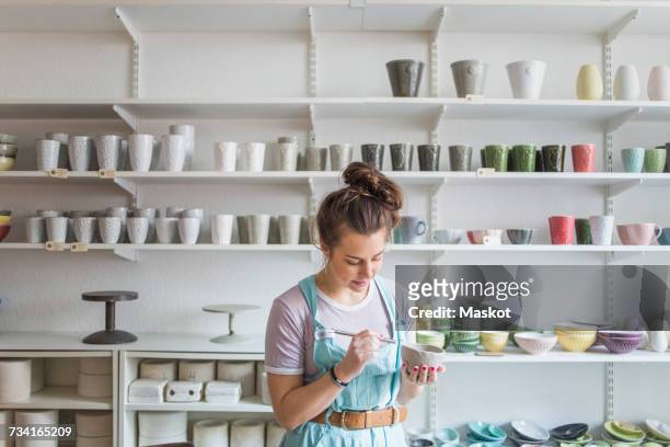 young female potter using hand tool on vase while standing against shelves at store - keramik stock-fotos und bilder