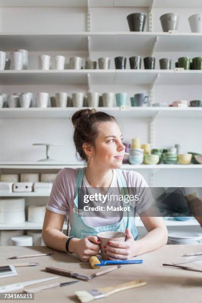 Young female potter sitting at workbench while looking away against shelves at store