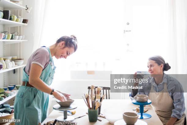 senior female potter looking at young ceramic artist molding clay against window in workshop - potter stock pictures, royalty-free photos & images