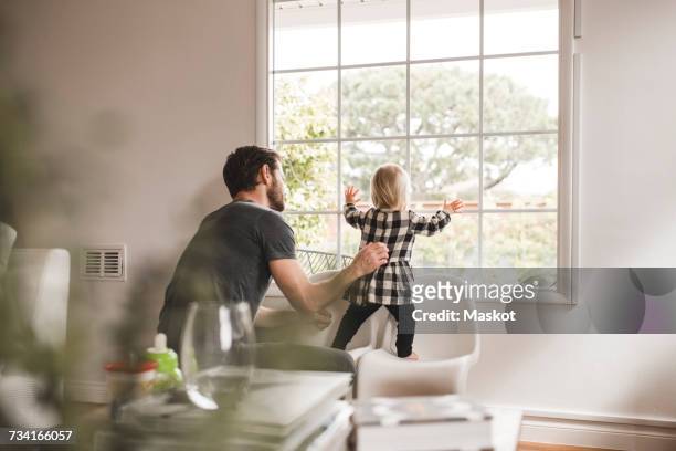 mid adult man with daughter looking through window at home - standing on chair stockfoto's en -beelden