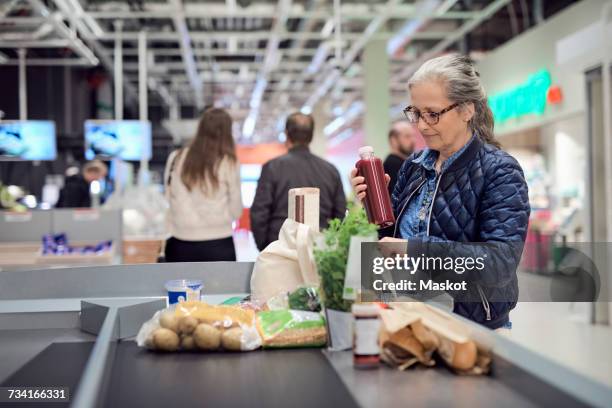 mature woman keeping juice bottle in bag while standing at checkout counter - kasse supermarkt stock-fotos und bilder