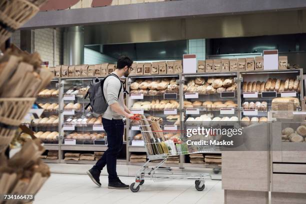 side view of man walking with cart while looking at breads in supermarket - pan fotografías e imágenes de stock