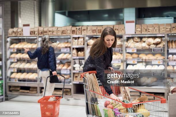 woman keeping bananas in shopping cart against rack at supermarket - shopping cart groceries stockfoto's en -beelden