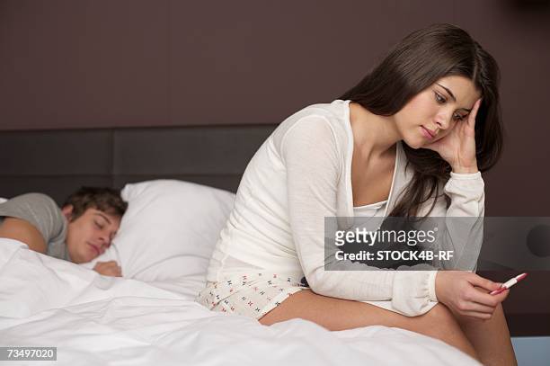 young brunette woman is sitting at the edge of a bed staring at a pregnancy test - onenightstand stock-fotos und bilder