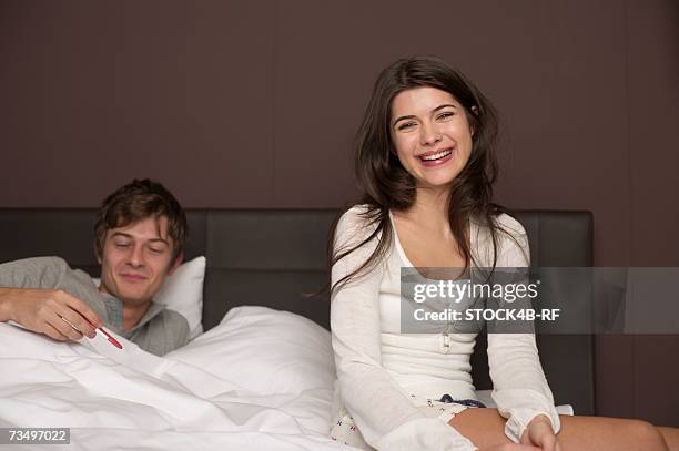 young brunette woman is sitting at the edge of a bed while her boyfriend is looking at a pregnancy test - onenightstand stock-fotos und bilder