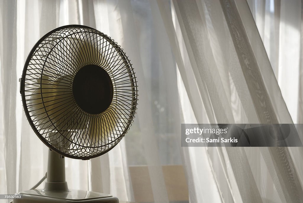 Electric fan beside apartment window with white curtains