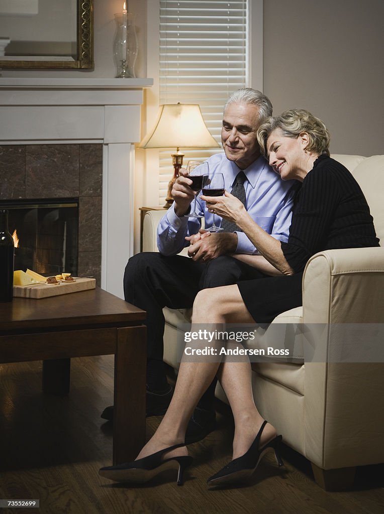 Senior couple sitting on sofa by fireplace, holding wine glasses, smiling
