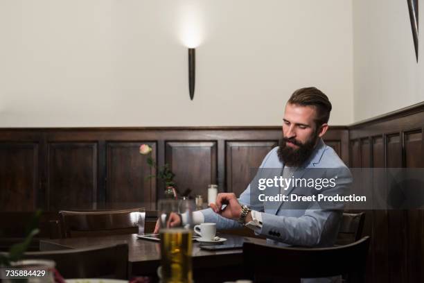 well-dressed man looking at watch while sitting at restaurant - newtechnology fotografías e imágenes de stock