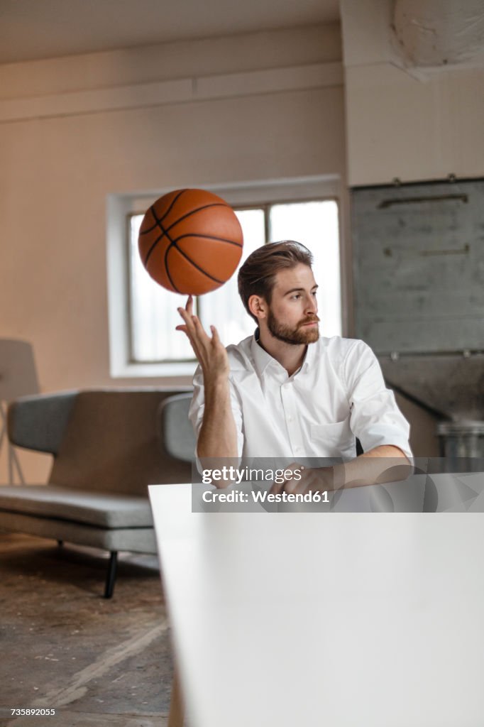 Portrait of pensive young businessman sitting at table in a loft balancing basketball on his finger