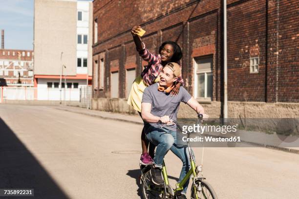 young couple riding bicycle in the street, woman standing on rack, taking selfies - business smartphone happy spring fotografías e imágenes de stock