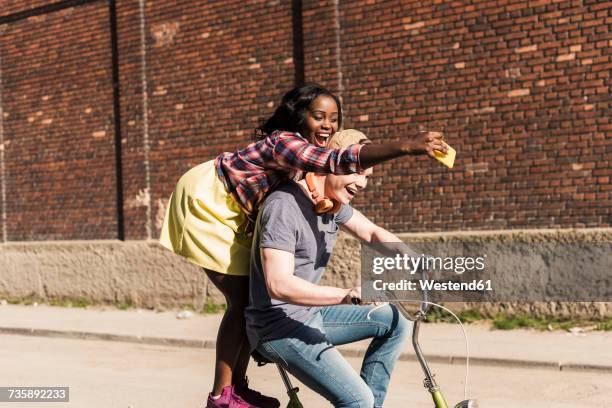 young couple riding bicycle in the street, woman standing on rack, taking selfies - gedeelde mobiliteit stockfoto's en -beelden