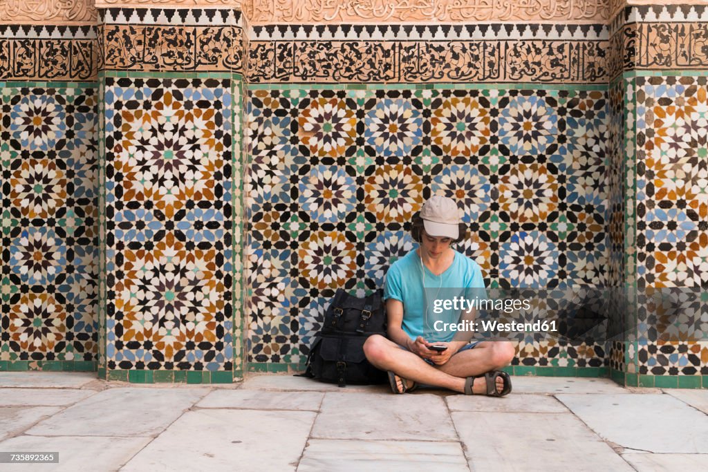 Morocco, Marrakesh, tourist sitting at tiled wall looking at cell phone