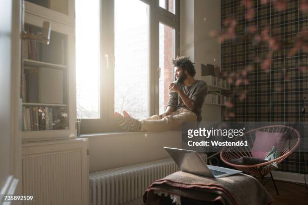 man sitting on window sill in living room looking outside holding a cup - coffee drink photos et images de collection