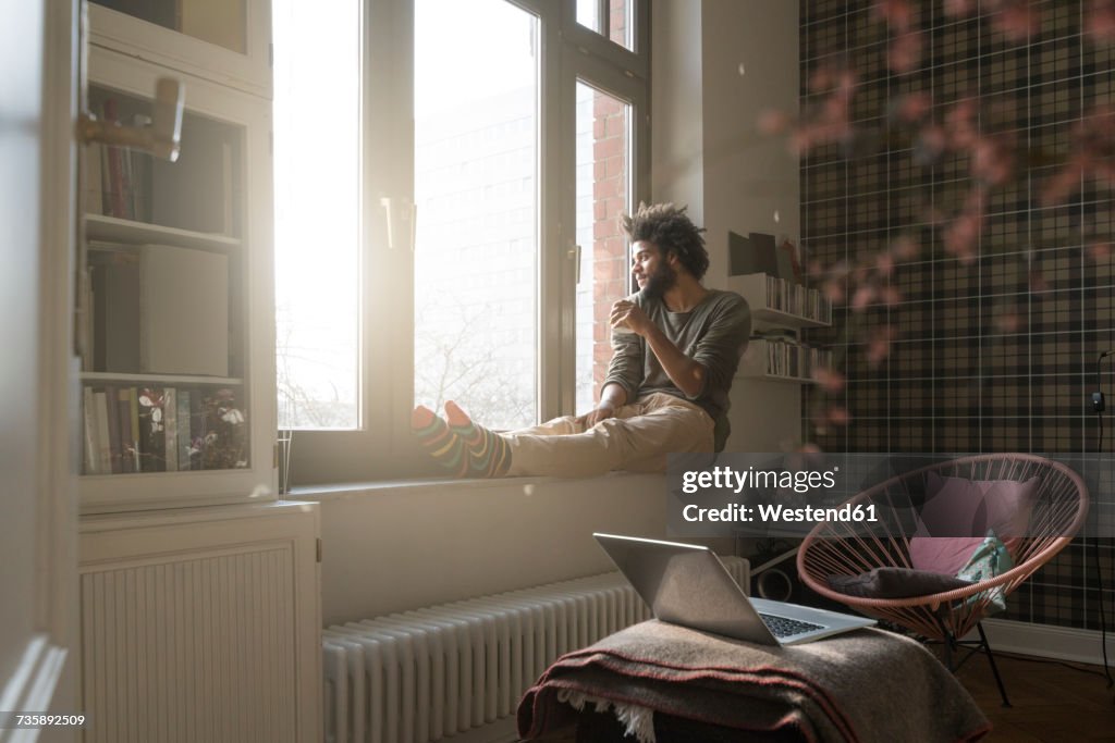 Man sitting on window sill in living room looking outside holding a cup