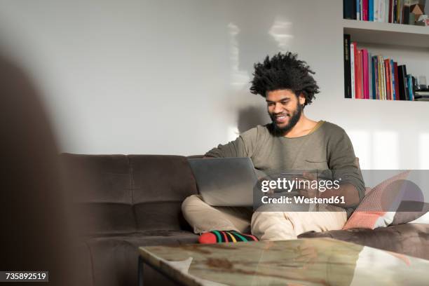 man sitting on couch in living room with laptop and credit card - shopping credit card stock-fotos und bilder
