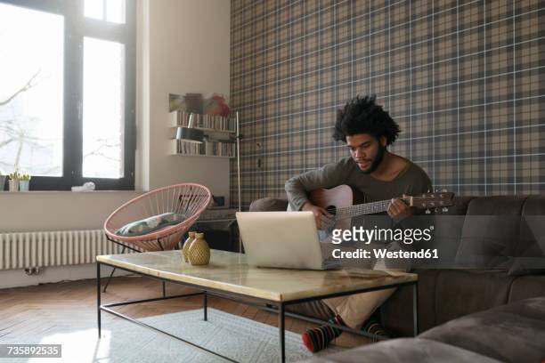man sitting in living room on sofa playing guitar in front of laptop - playing music photos et images de collection