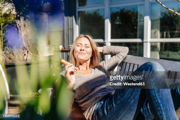 woman relaxing on garden bench eating a carrot - sunshine happy stock-fotos und bilder