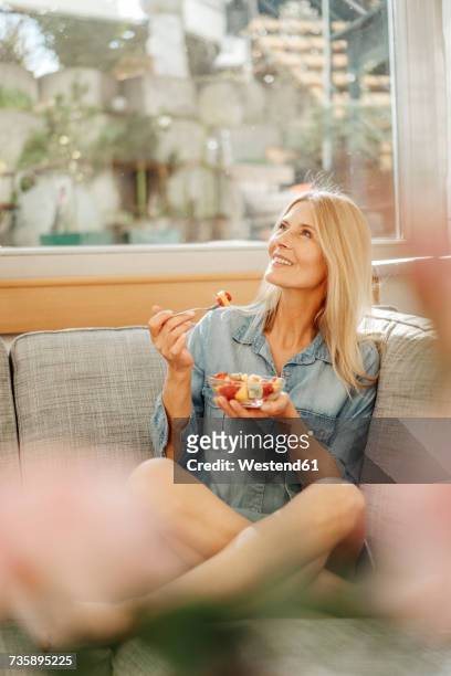 woman at home sitting on couch eating fruit salad - selective focus stock-fotos und bilder
