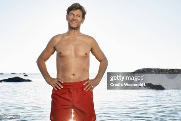 portrait of confident mature man wearing trunks standing at waterside - zwembroek stockfoto's en -beelden