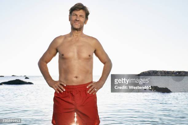 portrait of confident mature man wearing trunks standing at waterside - tronco nu imagens e fotografias de stock