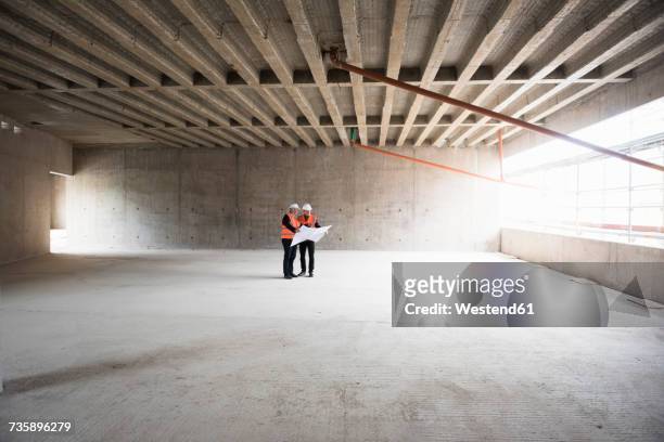 two men with plan wearing safety vests talking in building under construction - construction site stockfoto's en -beelden