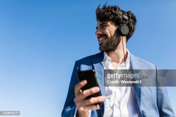 young businessman with smartphone wearing headphones under blue sky - business headphones stockfoto's en -beelden