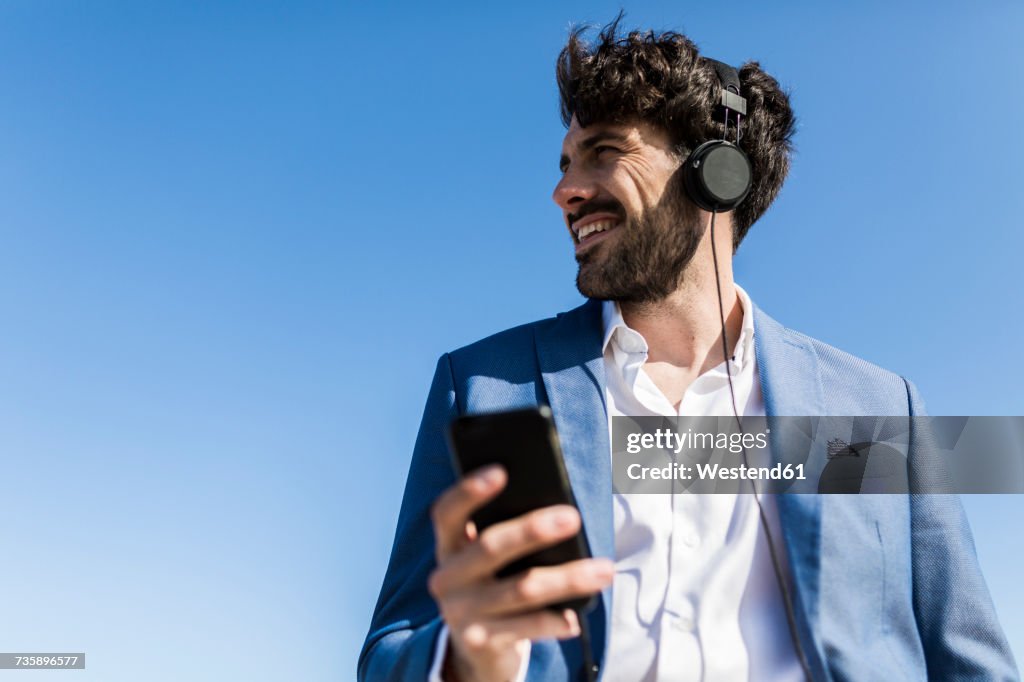 Young businessman with smartphone wearing headphones under blue sky