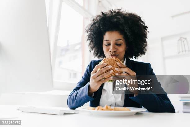 young businesswoman eating hamburger at her desk - woman junk food eating stockfoto's en -beelden