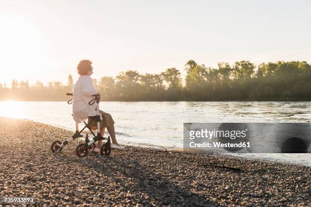 pensive senior woman sitting on her wheeled walker at riverside - rollator stock-fotos und bilder