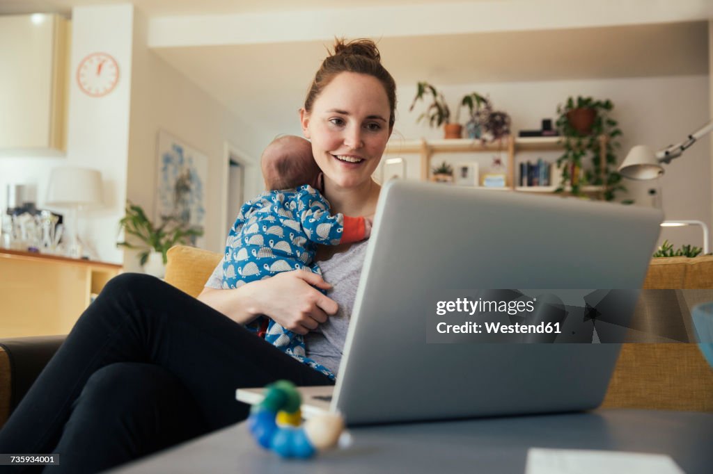 Mother using laptop and holding her newborn baby at home