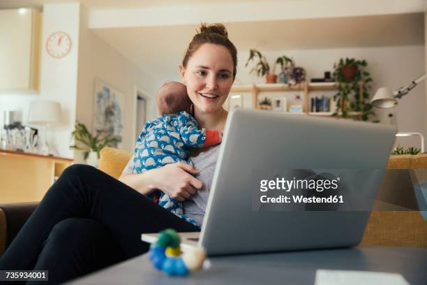 mother using laptop and holding her newborn baby at home - happy millennial at home photos et images de collection