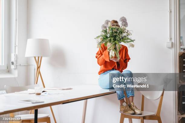 young woman sitting on table holding flower vase in front of her face - tímido fotografías e imágenes de stock