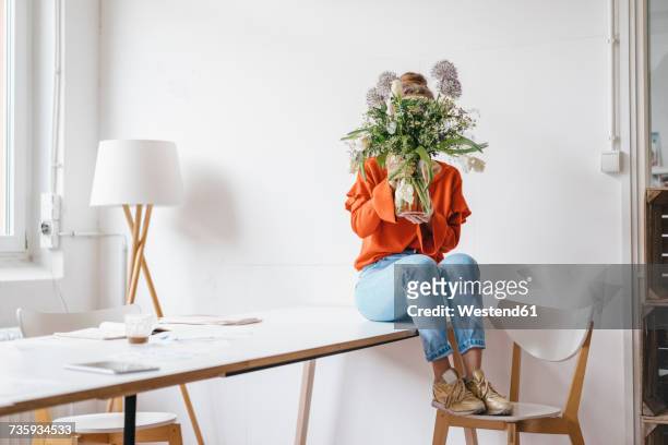 young woman sitting on table holding flower vase in front of her face - viso nascosto foto e immagini stock
